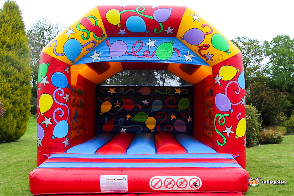 A colorful inflatable bouncy castle with balloons and stars design, set up on a grassy lawn, ready for children's entertainment.